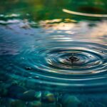 Minimalist image of a single droplet falling into still blue water, with soft ripples and gentle light symbolizing calm, connection, and the ripple effect of healing.